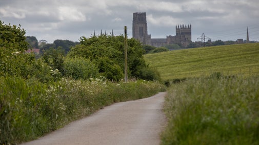 A narrow country lane between verges of lush summer vegetation in the foreground, leads towards hedgerows and trees in full leaf and a field of a bright yellow crop. Beyond, rooftops of the city's buildings sit under a cloudy but bright sky, dominated by Durham Cathedral.